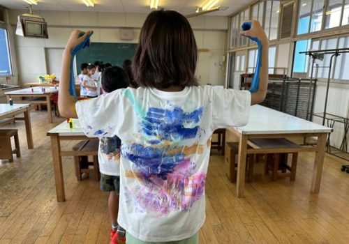 Strong mission & Vision - values, hands, hugs, a student holds up their arms showing solidarity with their classmates by showing off the shirt they are wearing that was painted by their classmates